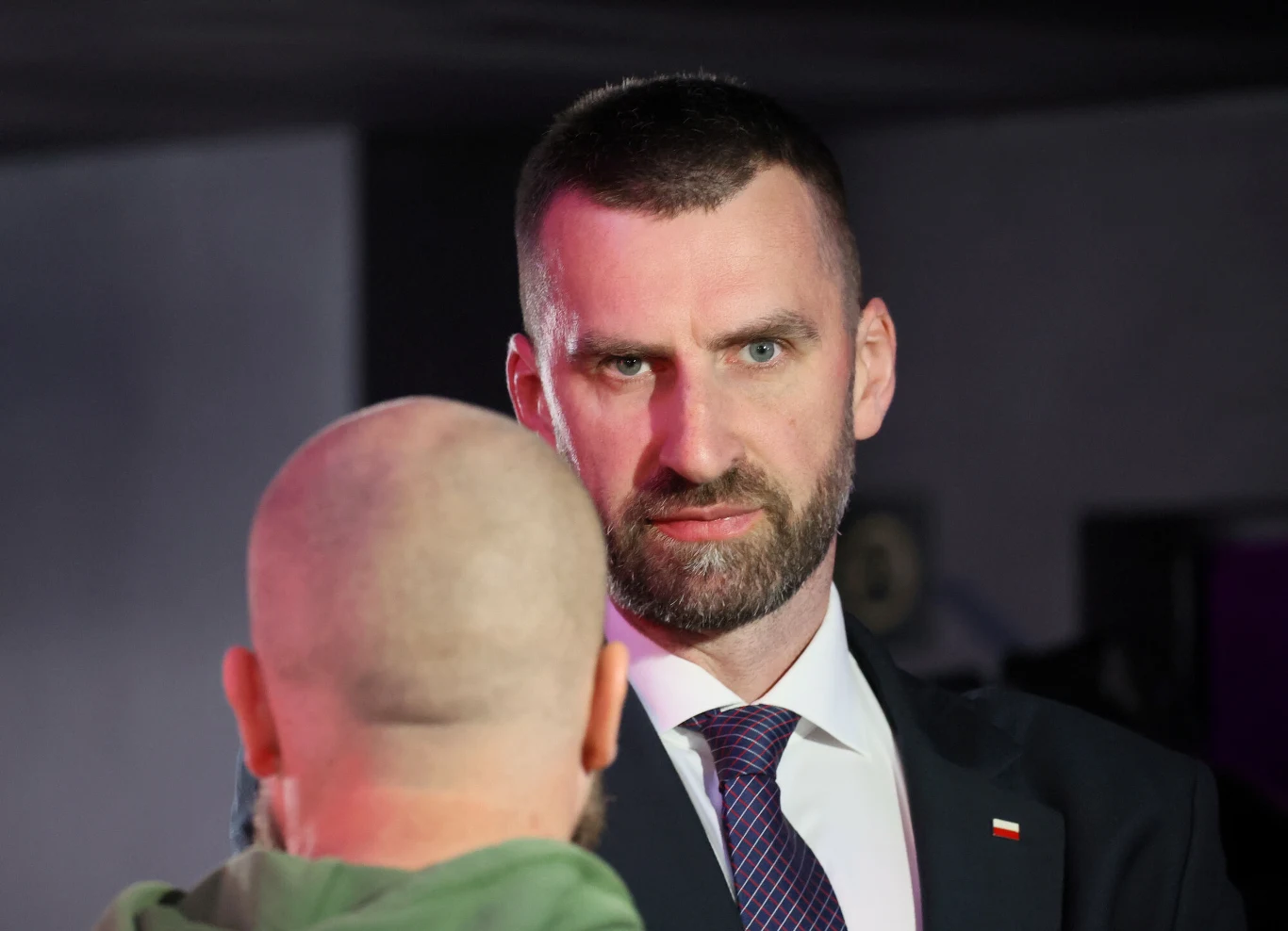 Marcin Możdżonek Mężczyzna w garniturze i krawacie, z poważnym wyrazem twarzy, patrzy na drugą osobę stojącą tyłem do obiektywu, która ma ogoloną głowę.