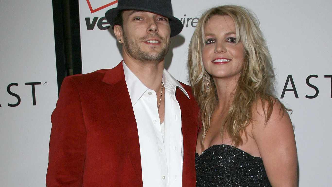 Kevin Federline i Britney Spears Dwie osoby stoją razem na tle ścianki eventowej, mężczyzna w czerwonej marynarce i białej koszuli, na głowie ma kapelusz, kobieta ma rozpuszczone blond włosy i błyszczącą czarną sukienkę, oboje uśmiechają się do aparatu.