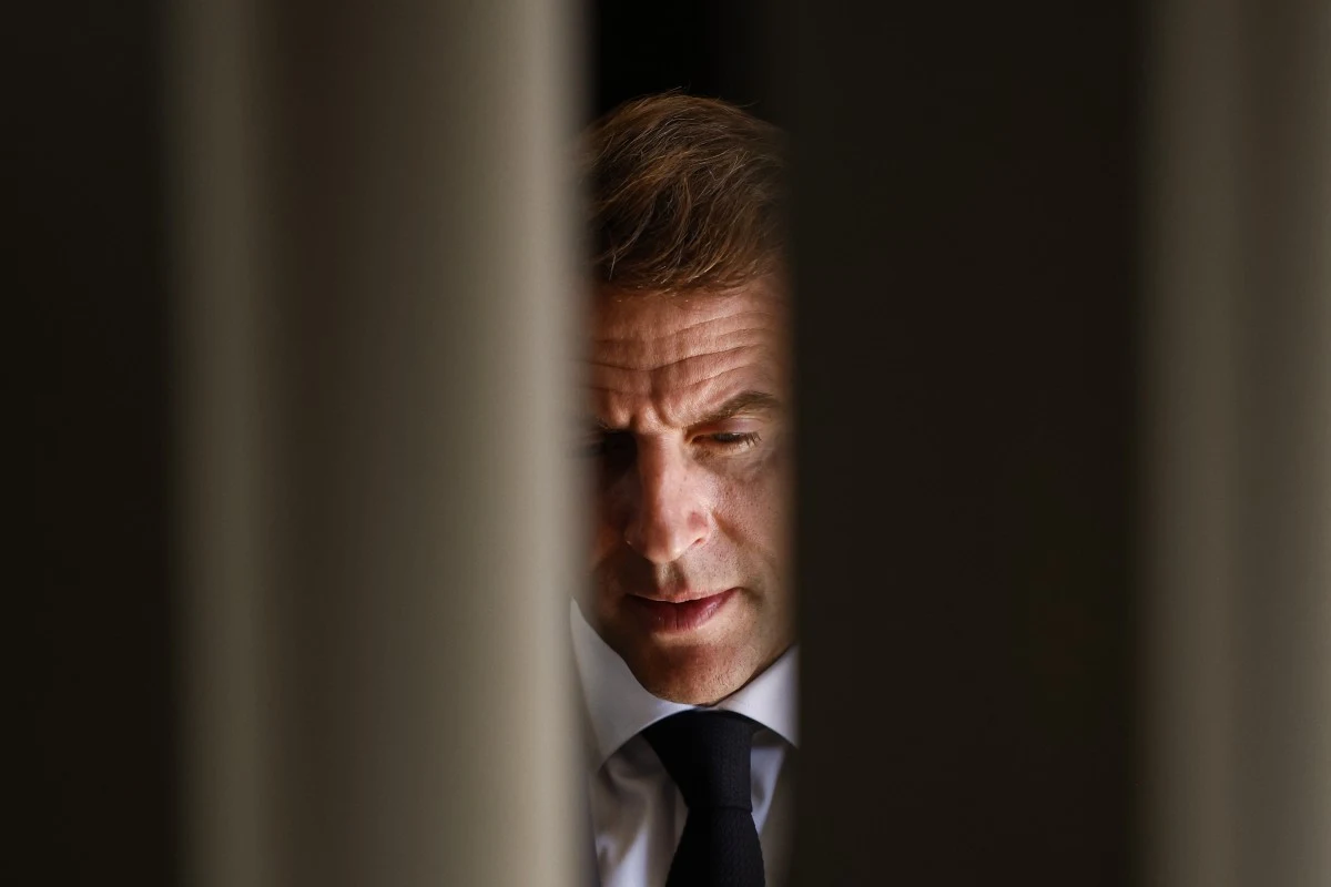 Emmanuel Macron rezygnuje z podwyższenia wieku emerytalnego Mężczyzna w garniturze uchwycony w półcieniu, widoczny przez szczeliny pomiędzy pionowymi elementami, skupiony wyraz twarzy podkreśla powagę sytuacji.