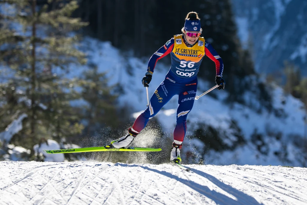 Flora Dolci Biegaczka narciarska w sportowym stroju i żółtym numerem startowym 56 podczas zawodów na śnieżnym stoku w zimowym krajobrazie, dynamiczna poza, promienie słońca podkreślają detale kombinezonu.