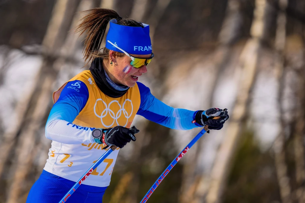 Flora Dolci Zawodniczka reprezentująca Francję podczas biegu narciarskiego na zimowych igrzyskach olimpijskich, ubrana w niebiesko-biały strój z numerem startowym, z widocznym malunkiem flagi Francji na policzku.