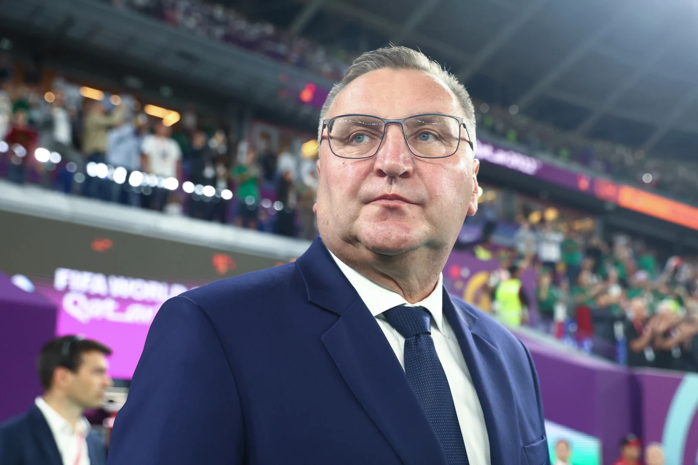 Mężczyzna w garniturze i okularach stoi na stadionie wśród tłumu kibiców, jest w centrum uwagi, w tle widać rozświetlone trybuny i osoby ubrane na sportowo.