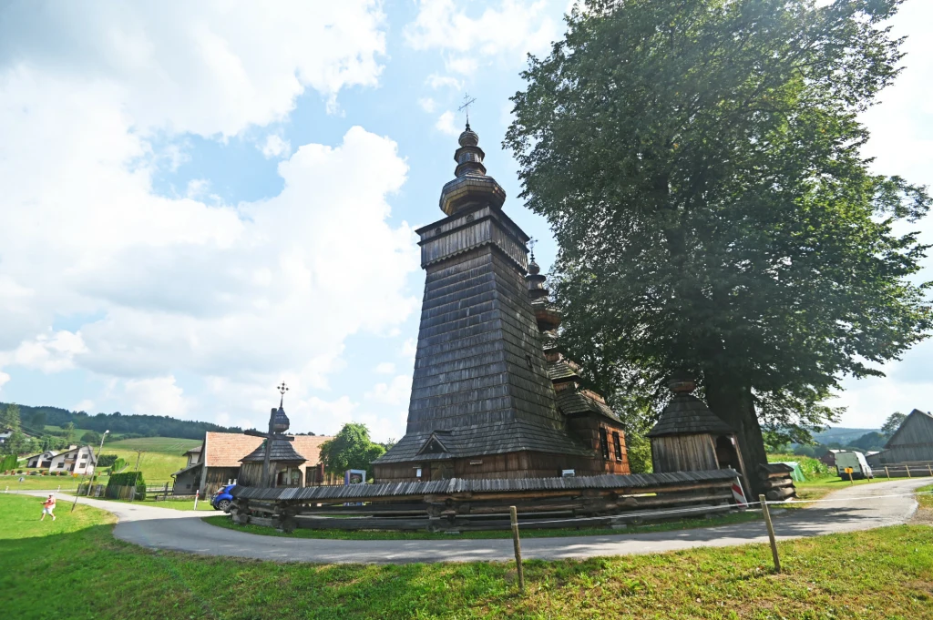 Drewniana cerkiew o charakterystycznej architekturze i wysokiej wieży, otoczona drewnianym ogrodzeniem, położona przy dużym drzewie, w tle zielone łąki i pojedyncze zabudowania wiejskie.