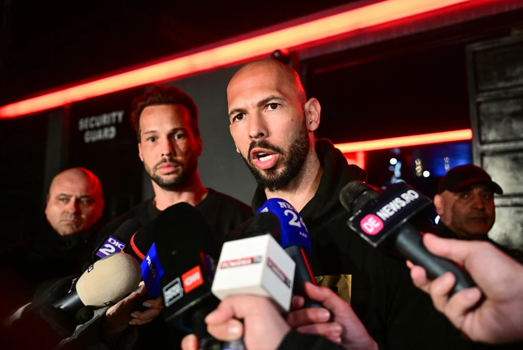 Andrew Tate Dwie osoby udzielają wywiadu otoczone mikrofonami różnych stacji telewizyjnych w ciemnym, zamkniętym pomieszczeniu, w tle widoczny ochroniarz oraz drzwi z napisem Security Guard.