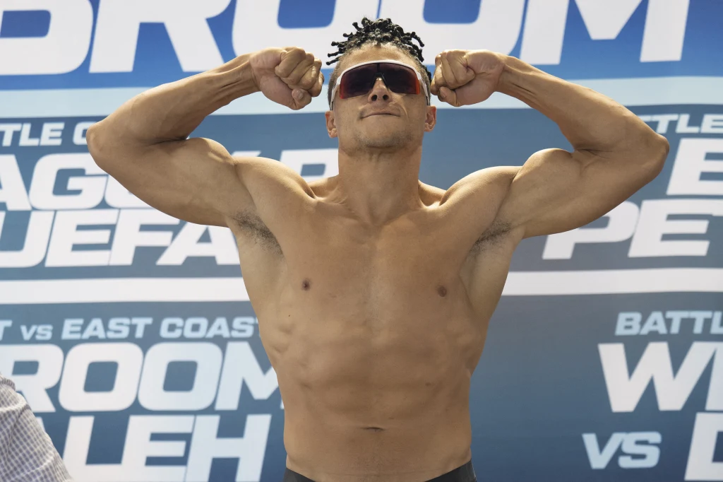 Chase DeMoor Mężczyzna w okularach przeciwsłonecznych prezentujący umięśnioną sylwetkę, z uniesionymi i zgiętymi w łokciach ramionami, na tle niebieskiej tablicy z napisami.