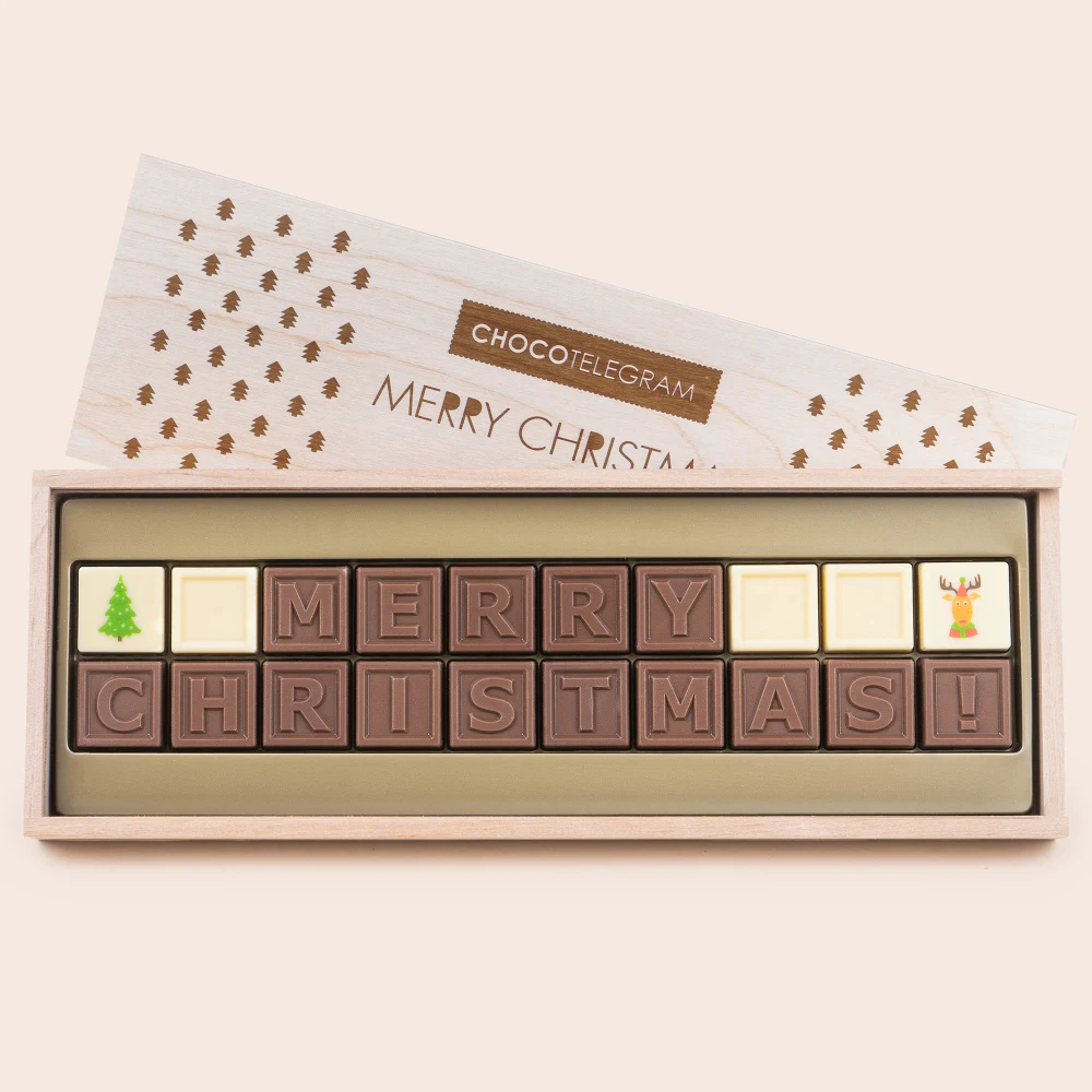 Czekolada od zawsze kojarzy się z przyjemnością Czekoladki ułożone w drewnianym pudełku tworzące napis Merry Christmas, z elementami ozdobnymi w postaci choinki i renifera na białych kostkach, opakowanie z motywem świątecznym.