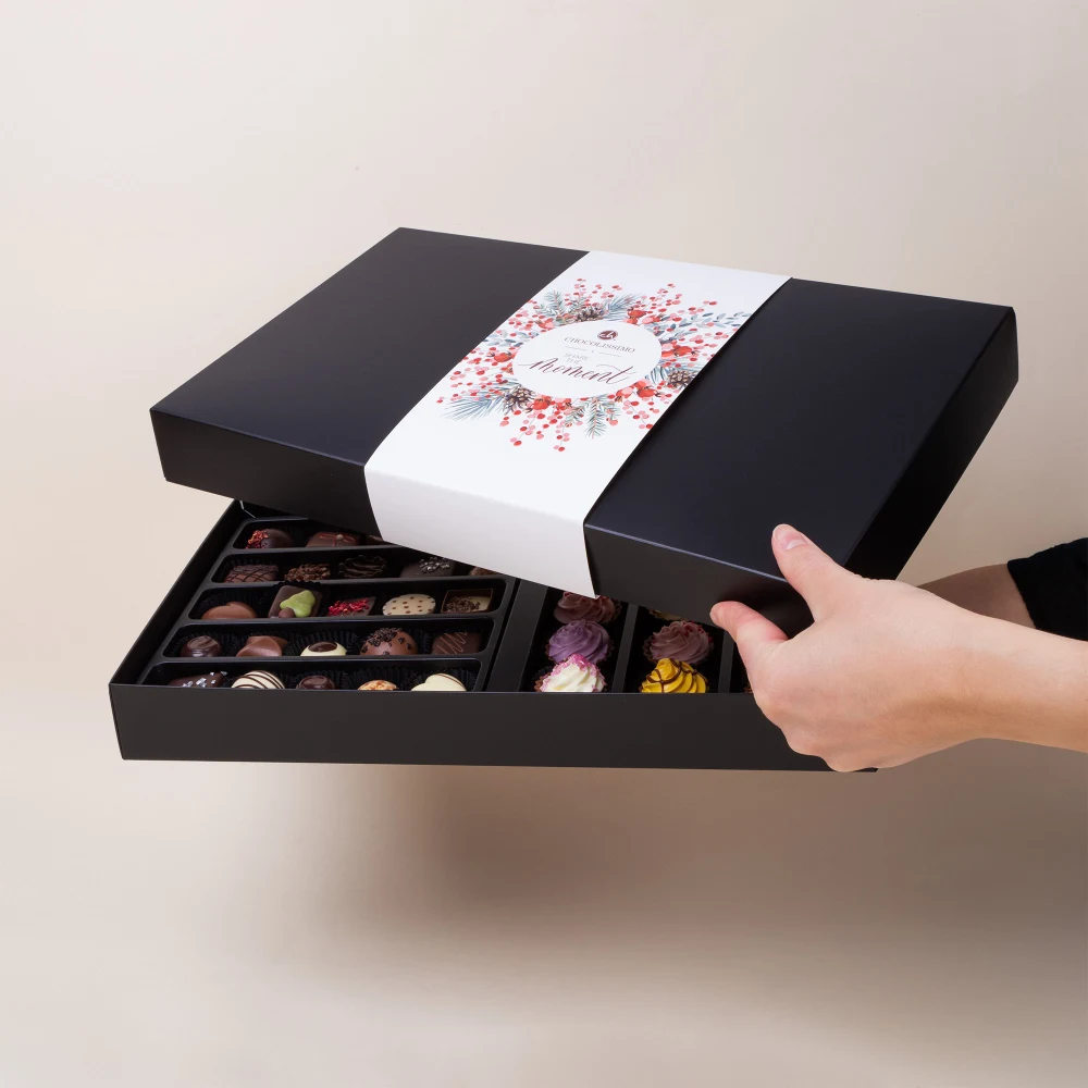 Otwarcie pudełka z pralinkami to mały rytuał Eleganckie czarne pudełko ozdobione białą banderolą z kwiatowym motywem, wypełnione różnorodnymi ręcznie robionymi pralinkami o różnych kształtach i kolorach, trzymane przez dwie dłonie na jasnym tle.