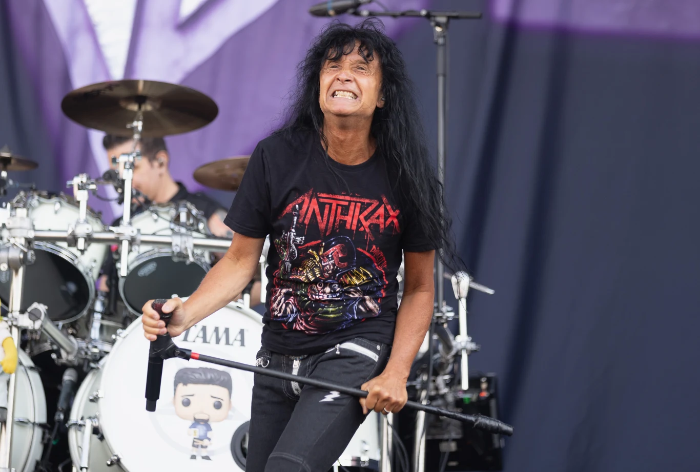 Joey Belladonna jest wokalistą grupy Anthrax Wokalista z długimi czarnymi włosami w koszulce zespołu Anthrax występujący na scenie podczas koncertu, z widocznym perkusistą i zestawem perkusyjnym w tle.