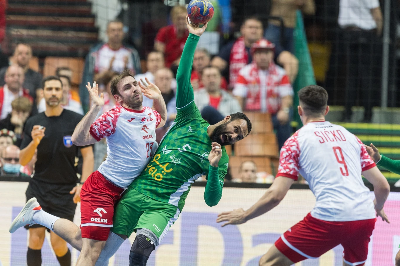 Arkadiusz Moryto i Szymon Sićko Zawodnik w zielonym stroju próbuje zdobyć punkt podczas meczu piłki ręcznej, będąc mocno blokowanym przez zawodnika drużyny przeciwnej w białych i czerwonych strojach. W tle widoczni są kolejni gracze oraz kibice na trybunach.