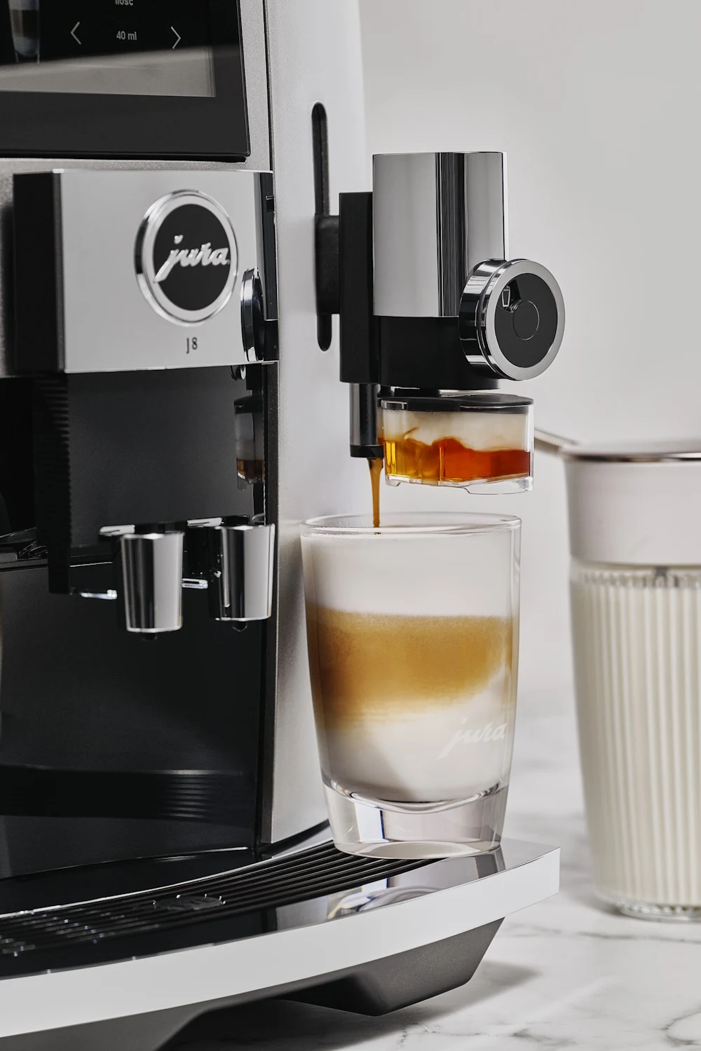 Piernikowe przyprawy w połączeniu z gorącą mleczną kawą rozgrzeją nas i otulą od środka Nowoczesny ekspres do kawy Jura podczas przygotowywania kawy latte, kawa delikatnie spływa do szklanki, w tle pojemnik ze spienionym mlekiem.