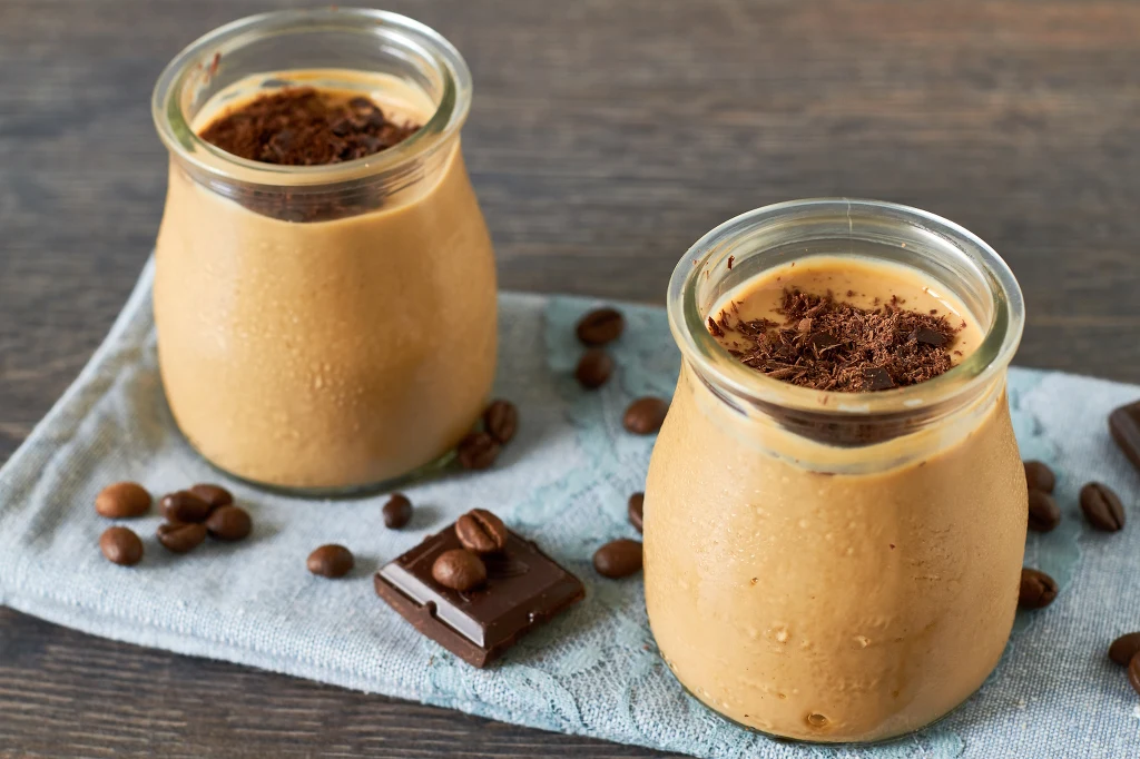 Alternatywą dla słodkich mlecznych kaw jest deser panna cotta z dodatkiem espresso Dwa szklane słoiczki wypełnione kremowym deserem kawowym, udekorowane wiórkami czekolady, ustawione na jasnej serwetce, wokół rozsypane ziarna kawy i kawałki gorzkiej czekolady.