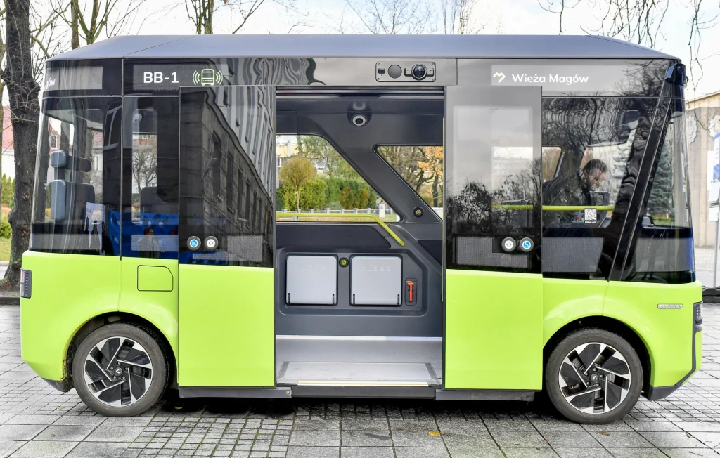 Autonomiczny elektryczny autobus o nowoczesnym designie, malowany na zielono-czarno, stoi zaparkowany na brukowanym chodniku; przez duże szyby widać wnętrze pojazdu oraz kierowcę.