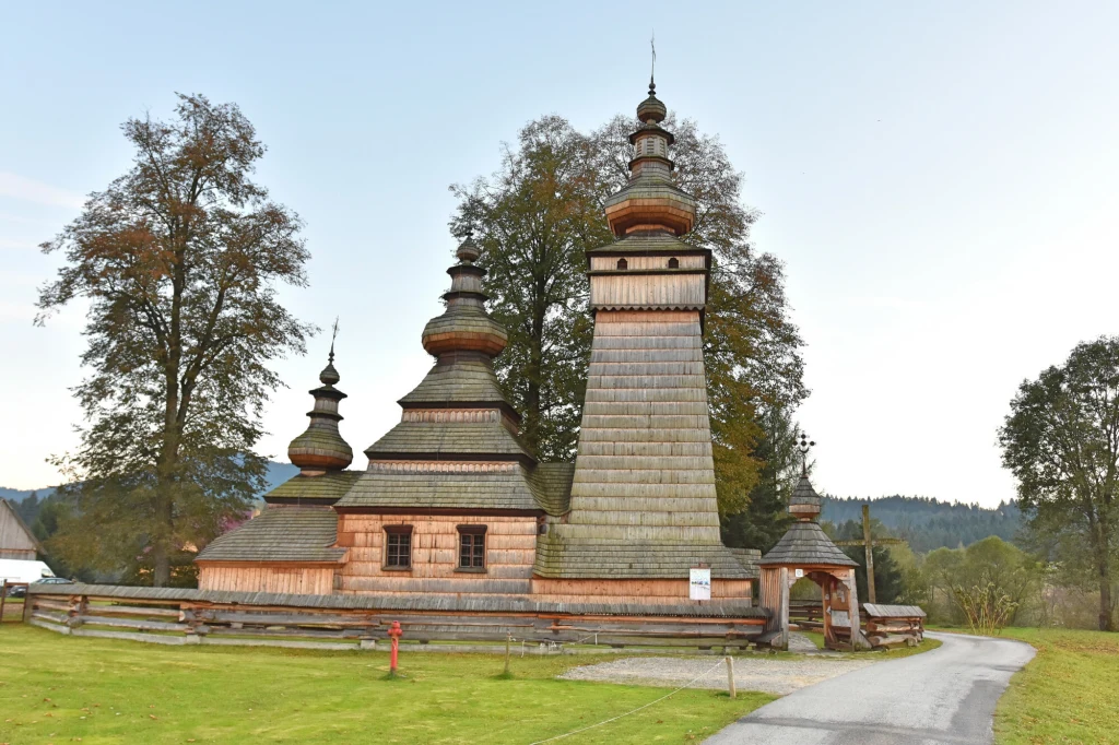 Drewniana cerkiew z trzema charakterystycznymi wieżami, otoczona niskim płotem i zielonym terenem, w tle drzewa i fragmenty gór, spokojna wiejska okolica.
