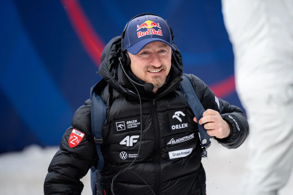 Mężczyzna w kurtce zimowej z licznymi naszywkami sponsorów, z mikrofonem na głowie i czapką z logo Red Bull, uśmiecha się podczas zimowego wydarzenia sportowego