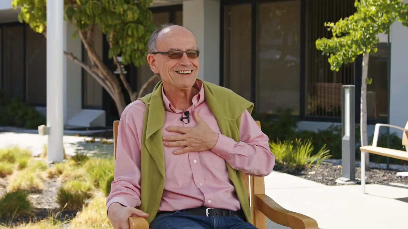 Thomas Sudhof podczas rozmowy z Maciejem Kaweckim Starszy mężczyzna w okularach siedzi na ławce na zewnątrz, ubrany w różową koszulę z oliwkowym swetrem przerzuconym przez ramiona, uśmiecha się szeroko i trzyma rękę na piersi, w tle roślinność i budynek z dużymi oknami.