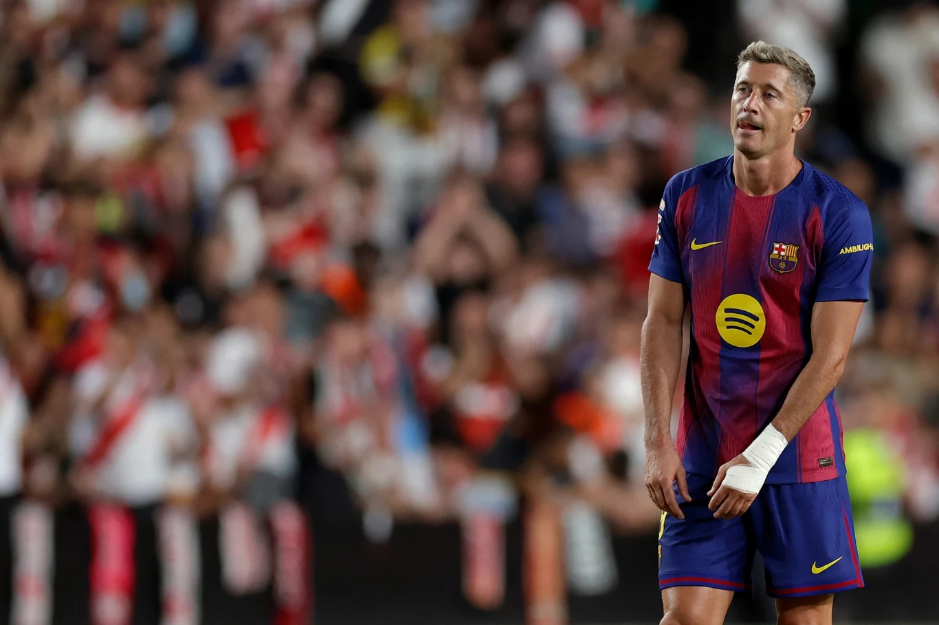 Robert Lewandowski Piłkarz w stroju Barcelony na boisku podczas meczu, z opatrunkiem na lewej ręce, w tle rozmyty tłum kibiców na trybunach.