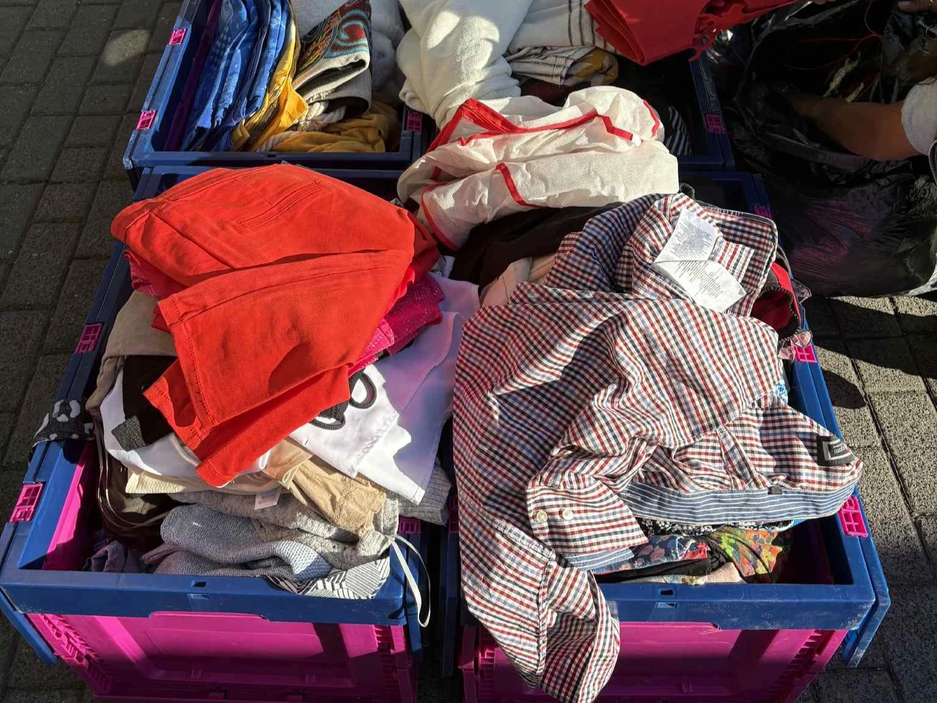 Stosy używanej odzieży w plastikowych pojemnikach o różnych kolorach, ustawione na chodniku w świetle słonecznym, ubrania w różnych kolorach, materiałach i wzorach