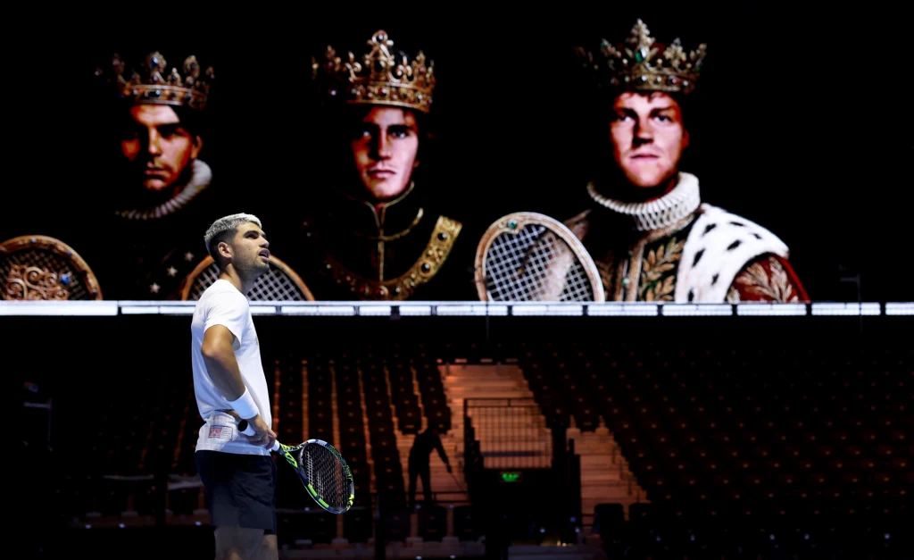 Carlos Alcaraz podczas turnieju Six Kings Slam Zawodnik tenisowy stojący na korcie z rakietą w ręku, na tle dużego ekranu z wizerunkami trzech mężczyzn ubranych w królewskie szaty i korony, trzymających rakiety tenisowe.