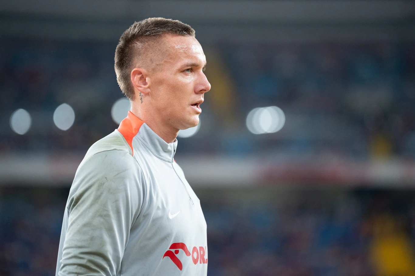 Piłkarz ubrany w jasną koszulkę sportową z widocznym logotypem, stoi na tle rozmytego stadionu, skupiony, z wyraźną fryzurą i tatuażem na uchu.