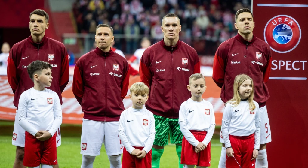 Polscy piłkarze w czerwonych kurtkach oraz dzieci w biało-czerwonych strojach stoją obok siebie na stadionie piłkarskim, w tle widać transparent z logo UEFA i kibiców na trybunach.
