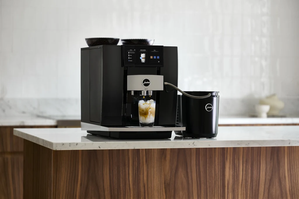 Codzienne czynności konserwacyjne wpisane są w cykl pracy ekspresów Jura Nowoczesny ekspres do kawy ustawiony na blacie kuchennym z kubkiem wypełnionym kawą oraz dołączonym pojemnikiem na mleko z logo producenta.