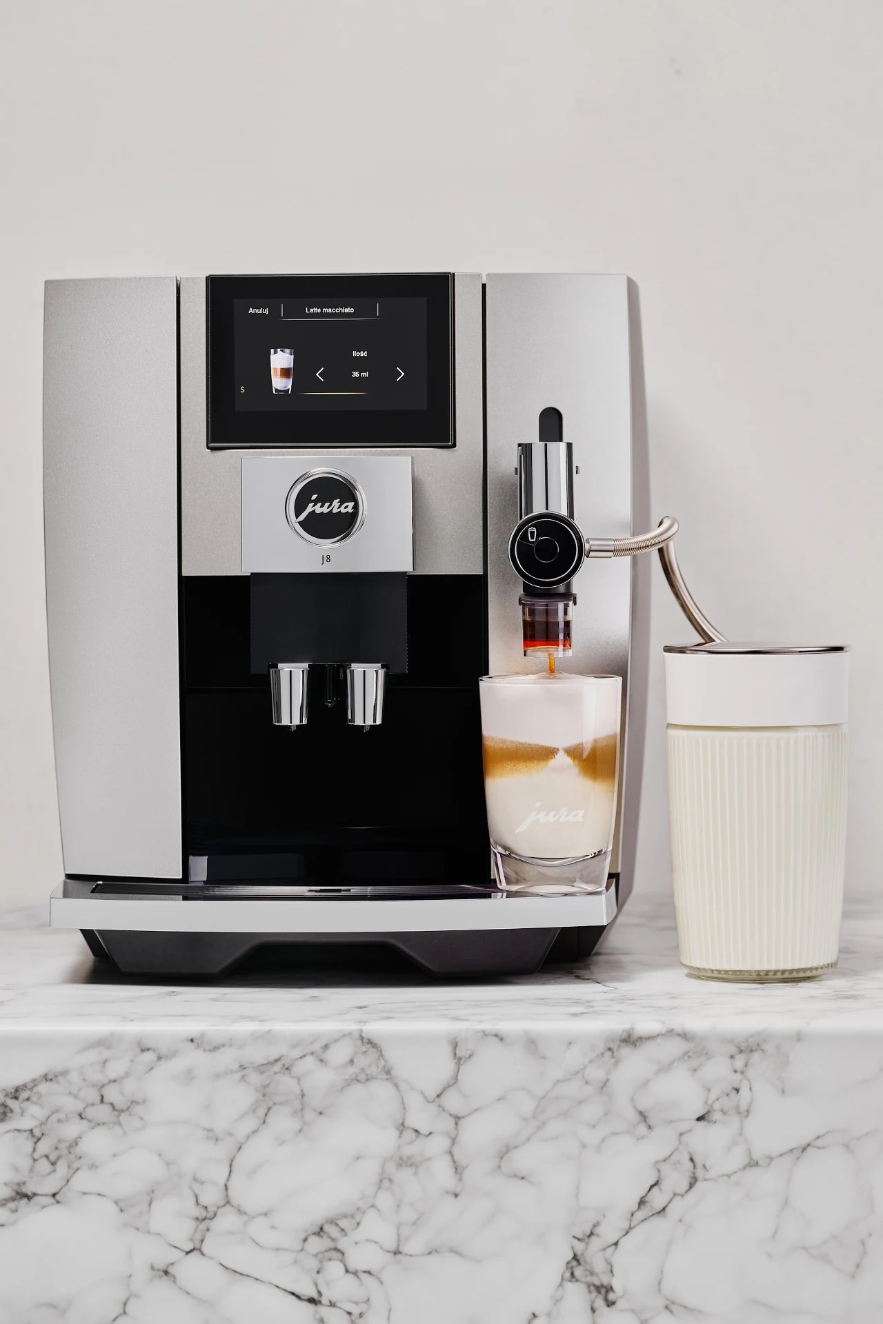 Czysty ekspres to nie luksus, ale fundament dobrej kawy Nowoczesny ekspres do kawy marki Jura przyrządza kawę z mleczną pianką do przezroczystej szklanki, obok stoi biały kubek na marmurowym blacie.