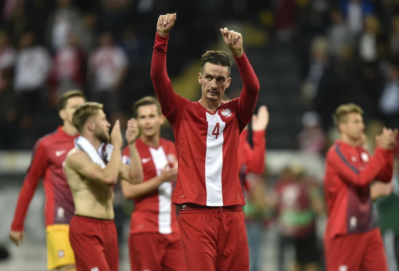 Piłkarz w czerwono-białym stroju z numerem 4 unosi w górę obie ręce w geście triumfu, w tle widoczni są inni zawodnicy drużyny, część z nich bez koszulek, wszyscy na stadionie po zakończonym meczu.