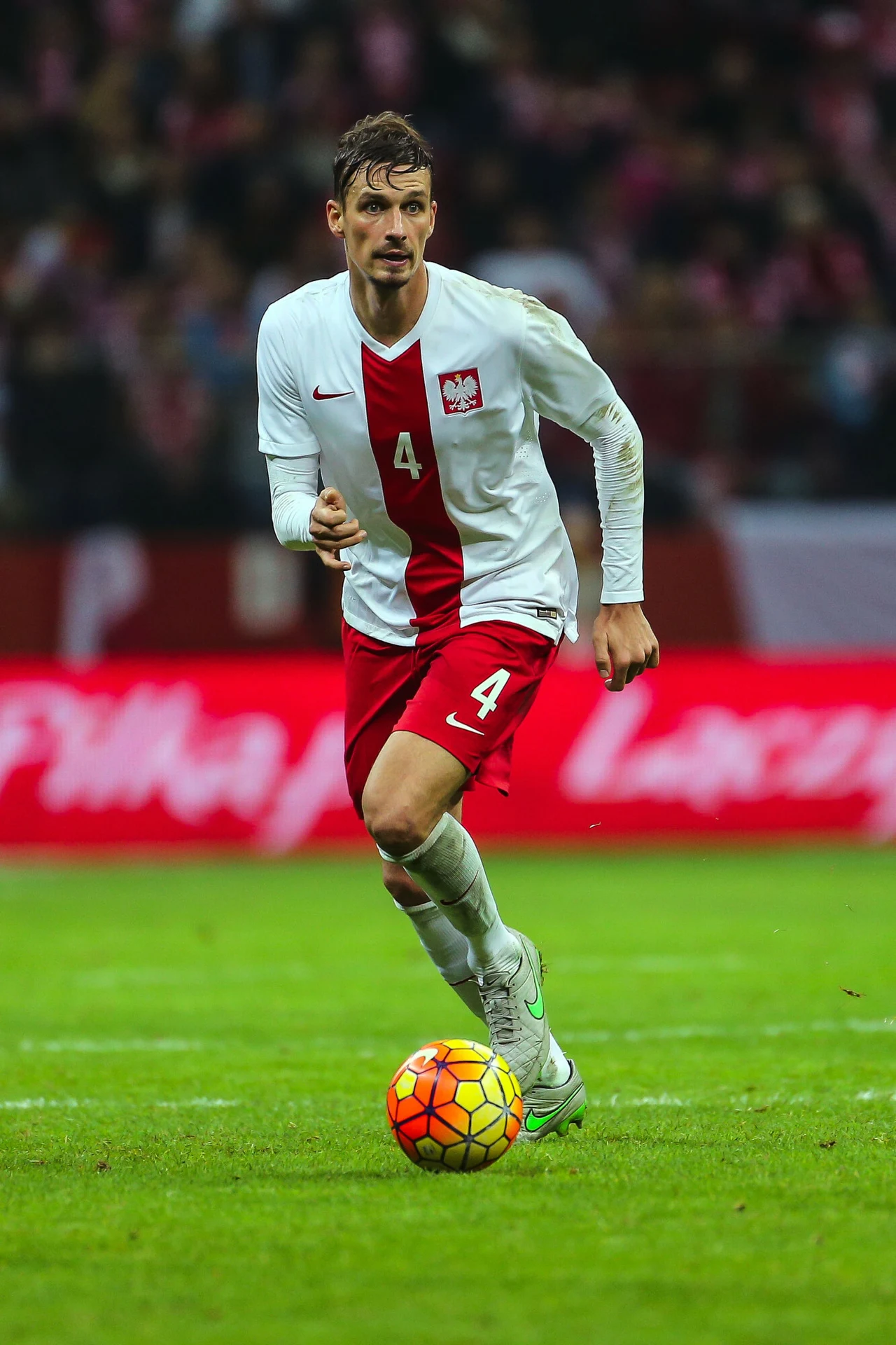 Piłkarz w białoczerwonym stroju z numerem 4 prowadzi piłkę podczas meczu na zielonej murawie stadionu, w tle widoczne trybuny wypełnione kibicami.