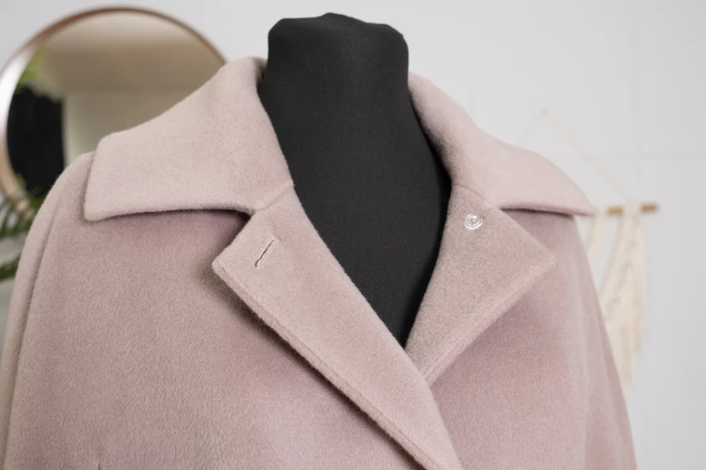 Jasnoróżowy, elegancki płaszcz z szerokim kołnierzem na czarnym manekinie, w tle widoczny fragment okrągłego lustra i jasnej ściany.