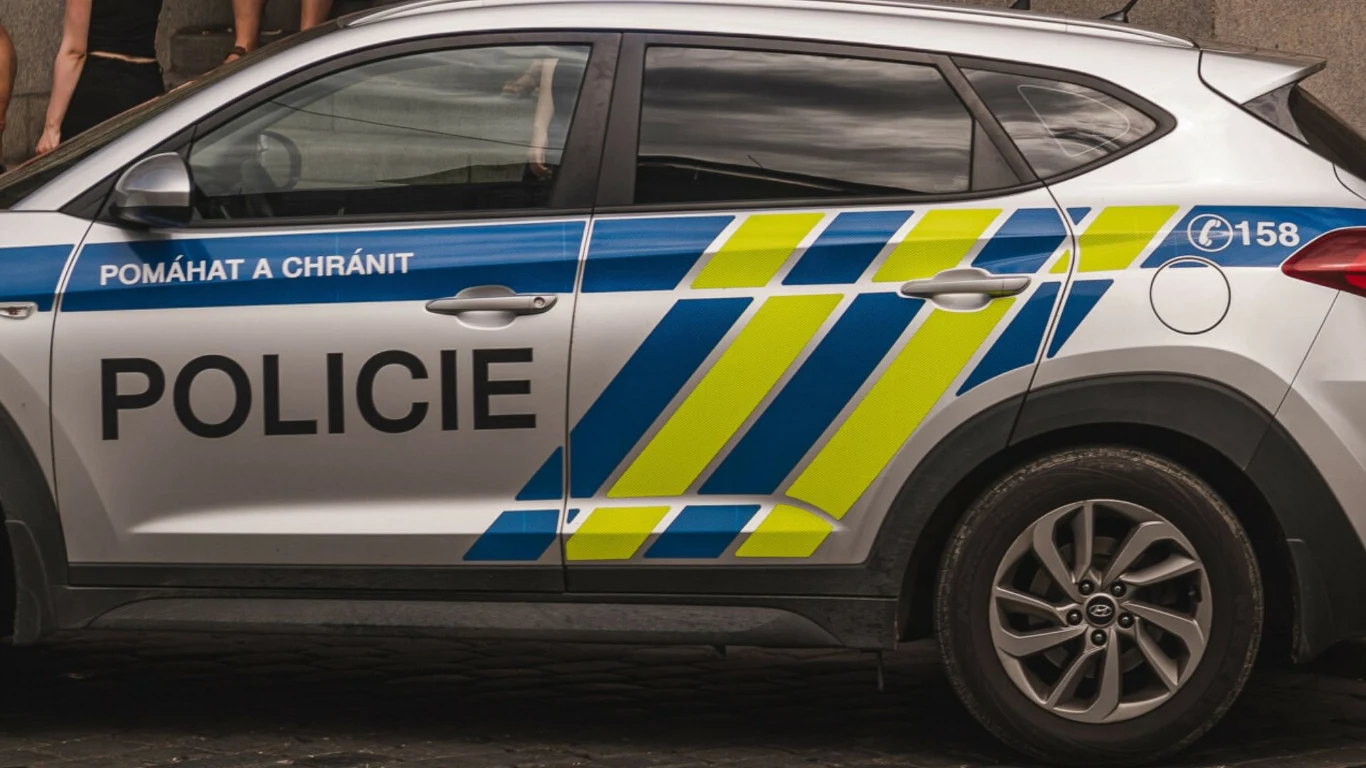 Czeska policja (zdj. ilustracyjne) Radiowóz policji z oznaczeniami żółto-niebieskimi i napisem 'Policie' na bocznych drzwiach, stojący na ulicy, widoczne częściowo koła oraz fragmenty nóg osób obok pojazdu.
