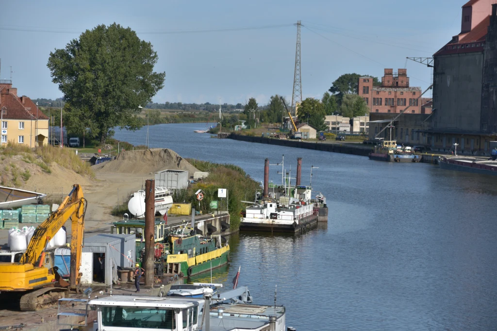 Rzeka z zacumowanymi barkami, na pierwszym planie sprzęt budowlany oraz dźwigi, w tle budynki przemysłowe i zieleń, po lewej stronie widoczne domy mieszkalne.