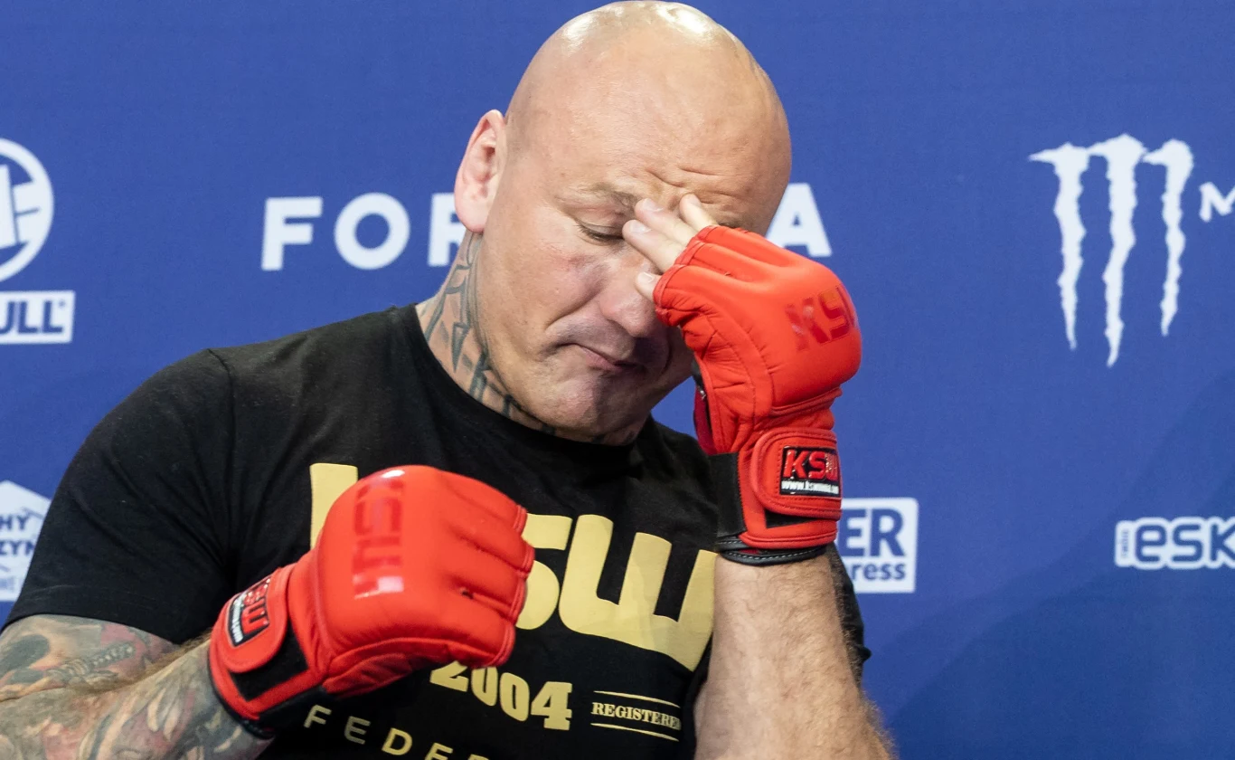 Zawodnik MMA w czerwonych rękawicach sportowych z charakterystycznymi tatuażami, zamyka oczy i trzyma się za twarz w geście zmęczenia lub frustracji na tle niebieskiej ścianki sponsorskiej.
