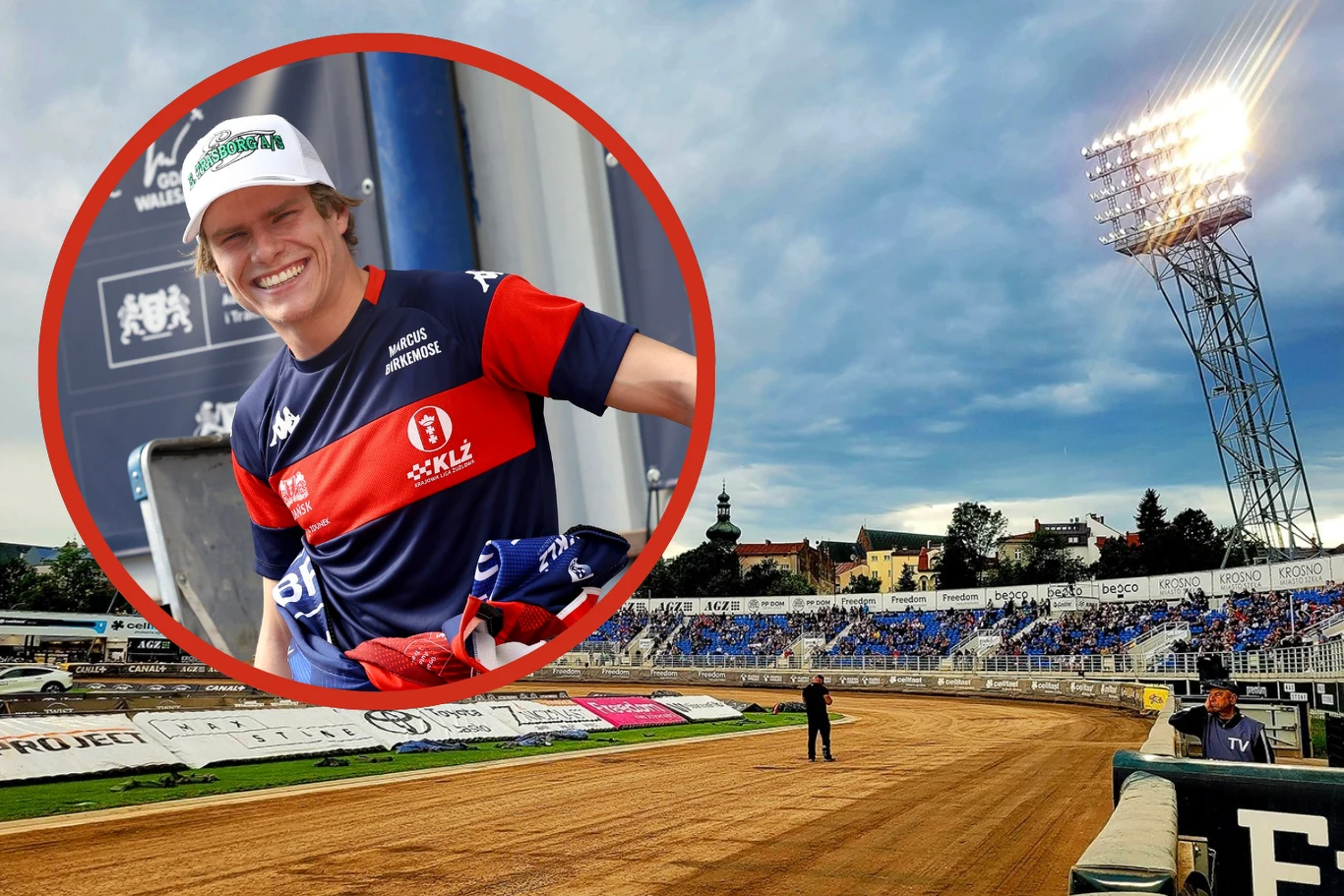 Sportowiec ubrany w strój klubowy żużlowy uśmiecha się na tle tablic sponsorskich, obok stadion żużlowy z trybunami, tor biegnący wokół widocznej murawy, w tle zabudowania miejskie.