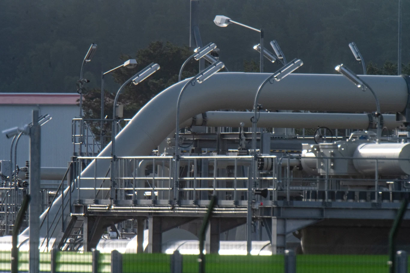 Na zdj. instalacje przesyłowe dla Nord Stream 2 na terenie Niemiec (stacja ciśnień/przepompownie gazu) Rurociągi gazowe i stalowe konstrukcje techniczne na terenie przemysłowym, otoczone barierkami i lampami oświetleniowymi, w tle drzewa oraz fragment budynku.