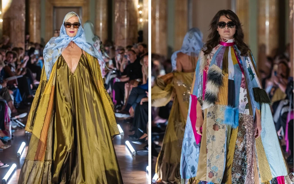 Modelki prezentują ekstrawaganckie, kolorowe kreacje na wybiegu, w tle widownia podczas pokazu mody w eleganckim, oświetlonym wnętrzu. Stroje składają się z luźnych, długich płaszczy i szali, a modelki mają ciemne okulary i nakrycia głowy.