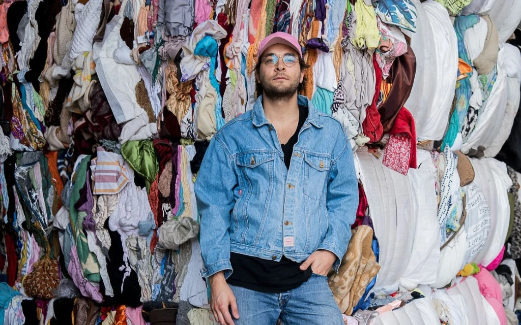 Mężczyzna w okularach i różowej czapce pozuje przed wielkim stosem kolorowych, używanych ubrań, ubrany w dżinsową kurtkę i dżinsy, lekko opiera się o ubrania.