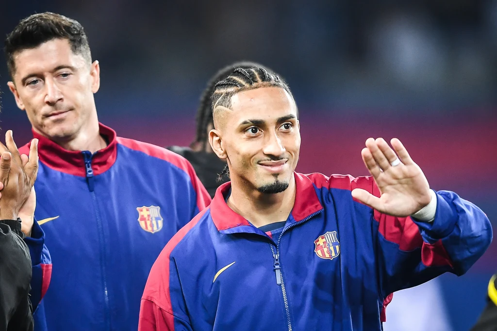 Dwóch piłkarzy drużyny FC Barcelona w oficjalnych dresach klubowych, jeden z nich uśmiecha się i macha ręką w kierunku obiektywu, w tle rozmyte sylwetki i trybuny stadionu.