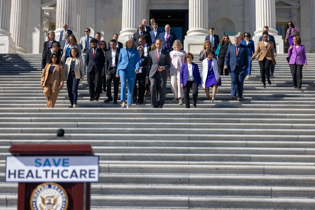 Demokraci z Izby Reprezentantów na schodach Kapitolu. W drodze na konferencję prasową na temat opieki zdrowotnej w 15. dniu zamknięcia rządu Grupa elegancko ubranych osób schodzi po schodach przed budynkiem o klasycznej architekturze z kolumnami, na pierwszym planie widoczna mównica z napisem Save Healthcare.
