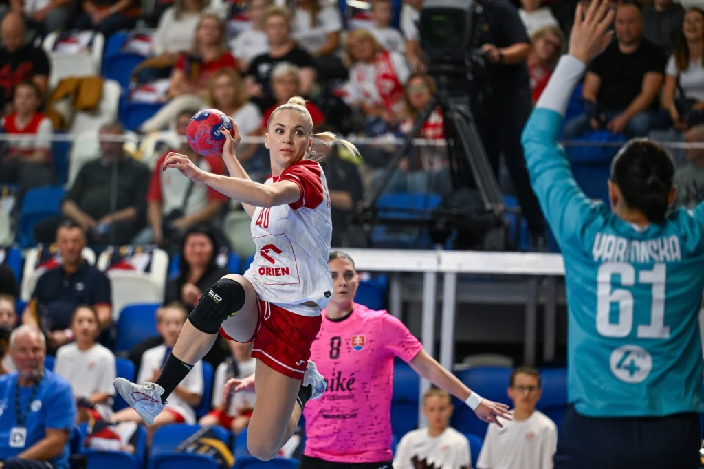 Zawodniczka w biało-czerwonym stroju unosi się w powietrzu podczas akcji rzutowej w hali sportowej, w tle widać bramkarkę drużyny przeciwnej w niebieskim stroju oraz kibiców na trybunach.