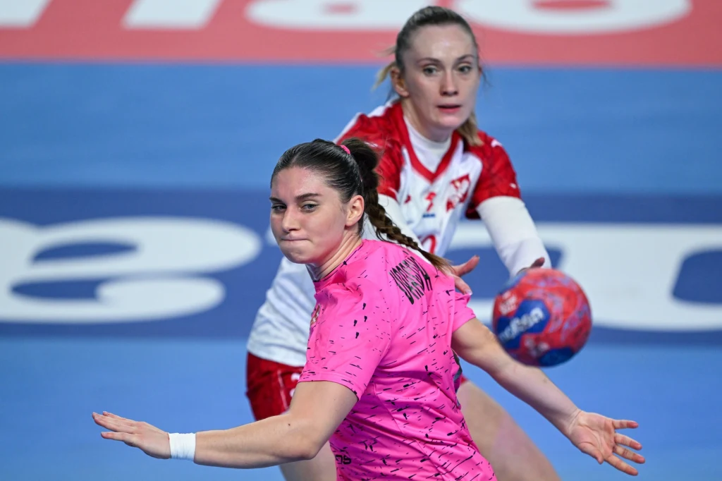 Dwie zawodniczki piłki ręcznej w trakcie dynamicznej akcji na boisku, jedna ubrana w różowy strój z piłką w ręku, druga w biało-czerwonym stroju w pozycji obronnej.