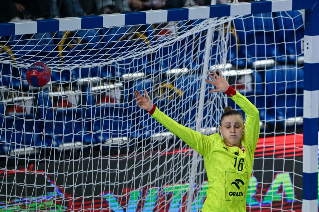 Bramkarka w żółtym stroju unosi ręce w geście obrony, przed nią piłka leci w stronę bramki, tło stanowią trybuny oraz bramka z siatką.