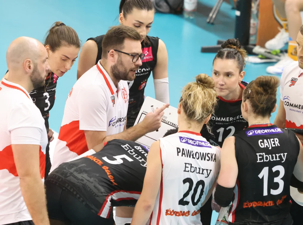 Trener drużyny siatkarskiej omawia strategię z zawodniczkami podczas przerwy w meczu, zawodniczki w skupieniu słuchają wskazówek, ubrane w stroje sportowe z widocznymi nazwiskami oraz nazwami sponsorów.