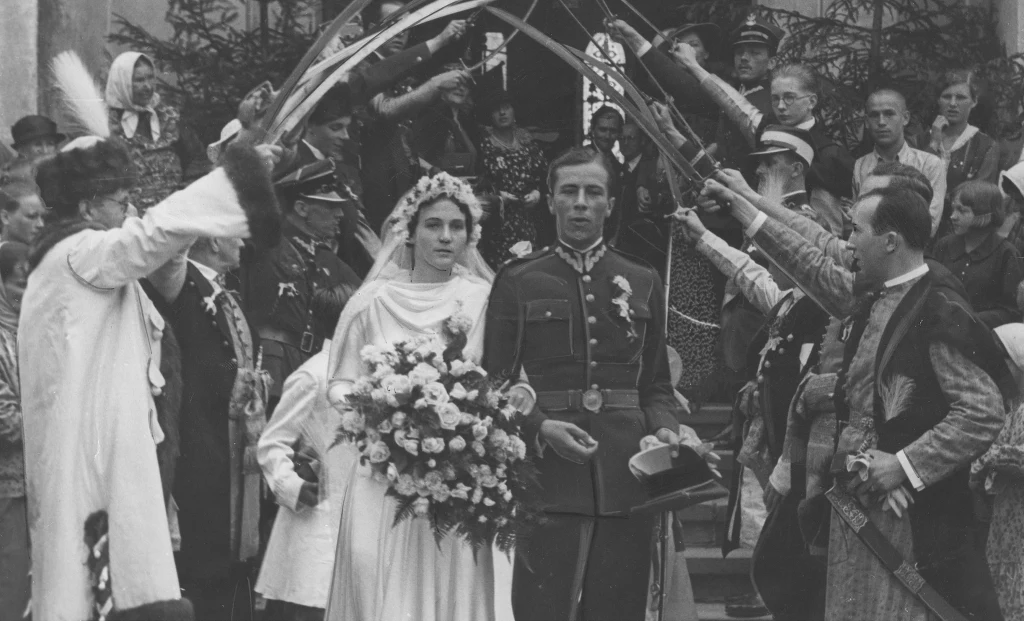 Ślub księcia Adama Michała Józefa Czartoryskiego z hrabianką Jadwigą Stadnicką w 1937 roku Para młoda wychodząca z kościoła w towarzystwie żołnierzy, panna młoda z bukietem kwiatów w białej sukni, pan młody w mundurze wojskowym, nad ich głowami żołnierze tworzą szpaler z szabli, wokół tłum gości ubranych w tradycyjne i wojskowe stroje.