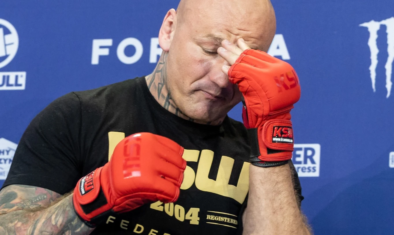 Zawodnik MMA w czerwonych rękawicach z logo organizacji KSW zasłania twarz dłonią, jakby wyrażał zmęczenie lub rozczarowanie, w tle widoczne są logotypy sponsorów i niebieska ścianka prasowa.
