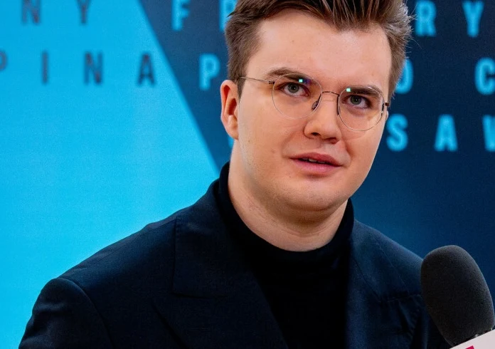 Piotr Alexewicz zagra w finale Konkursu Chopinowskiego Młody mężczyzna z okularami i starannie ułożonymi włosami, ubrany w ciemną marynarkę i czarny golf, trzyma mikrofon, tło w odcieniach niebieskiego z rozmazanym napisem.
