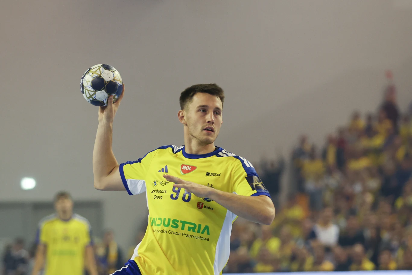 Piotr Jarosiewicz (Industria Kielce) Zawodnik piłki ręcznej w żółtym stroju drużyny podczas meczu szykuje się do rzutu piłką, w tle widoczna publiczność i inni gracze.