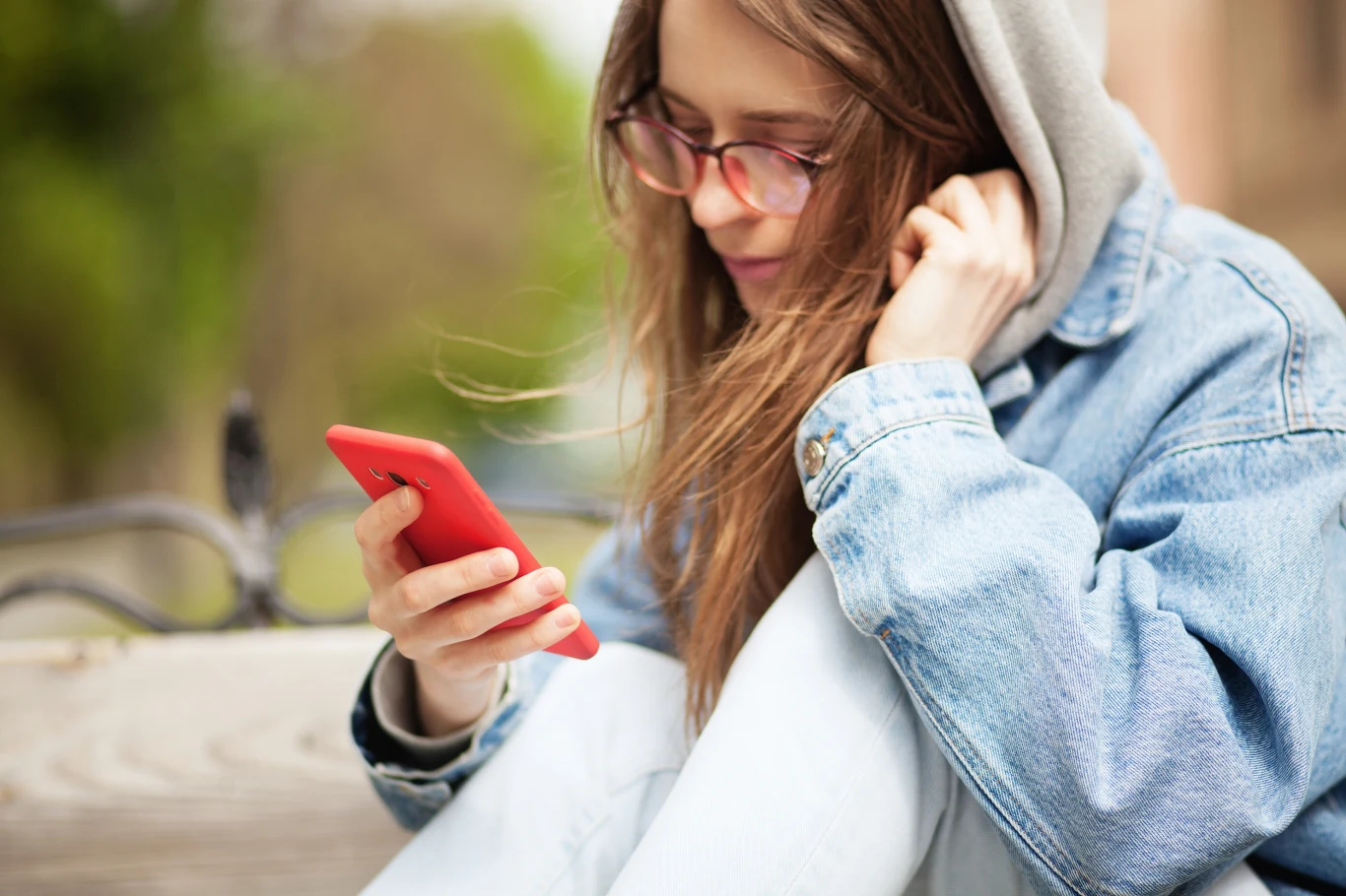 Około połowa nastolatków, bo 48 procent badanych, twierdzi, że media społecznościowe mają głównie negatywny wpływ na osoby w ich wieku. Młoda kobieta w okularach i dżinsowej kurtce, ubrana na luzie, skupiona na czerwonym smartfonie, siedzi na zewnątrz na ławce z widokiem na zielone otoczenie.