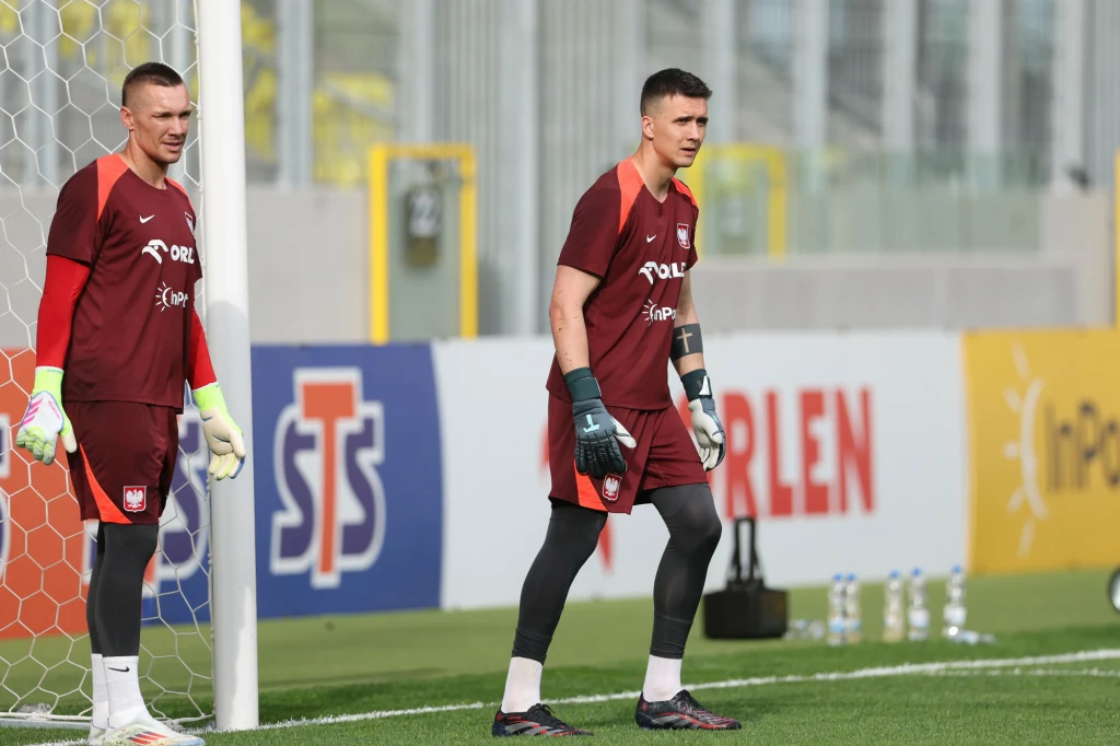 Dwóch bramkarzy ubranych w bordowe stroje treningowe stoi na murawie boiska, obok bramki, na tle reklam sponsorów widocznych na bandach.