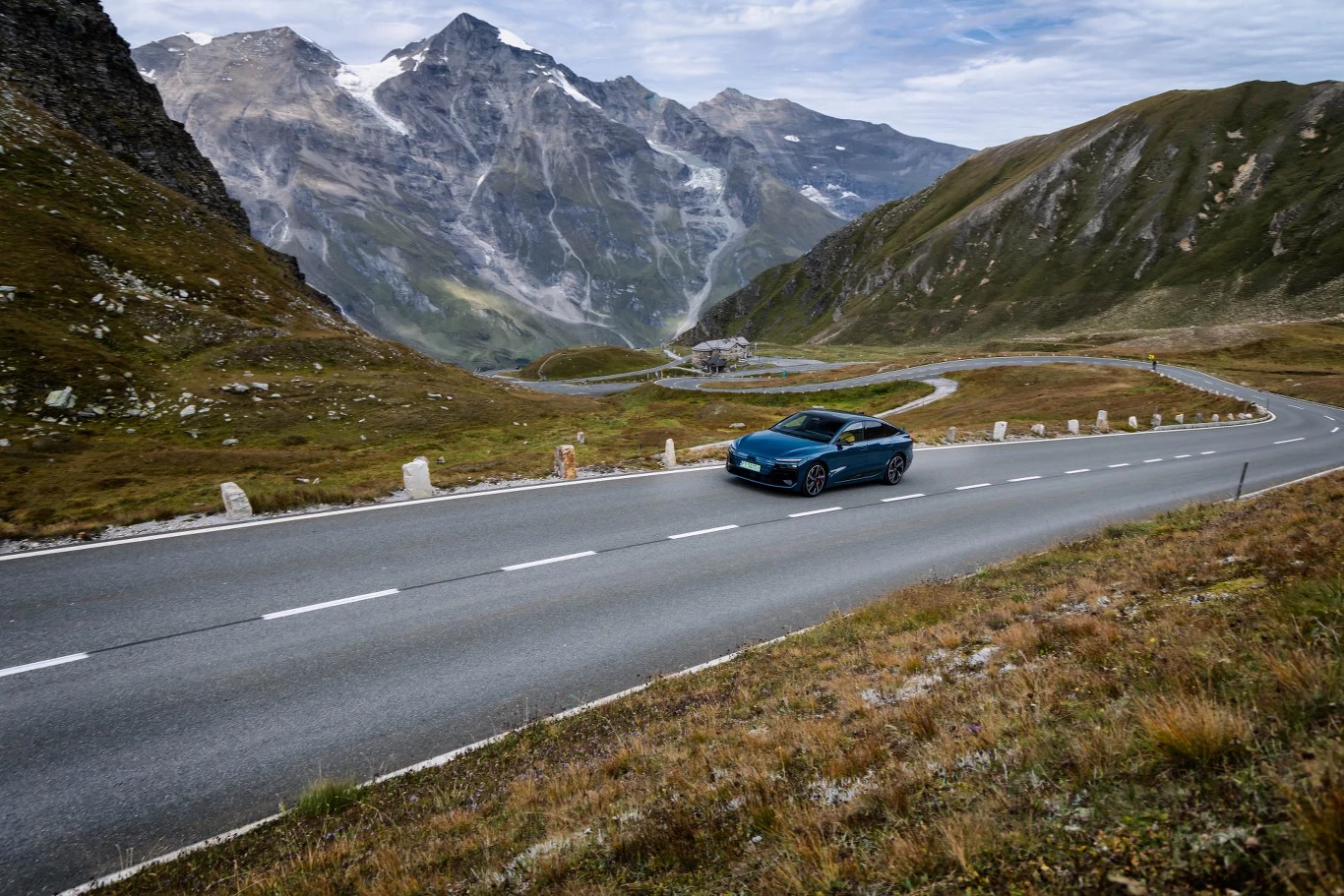 Audi S6 e-tron na Grossglockner Hochalpenstrasse. Elektryczna limuzyna w swoim żywiole – cisza, precyzja i 490 KM wśród alpejskich szczytów Samochód osobowy jedzie po krętej, górskiej drodze otoczonej zielonymi zboczami i wysokimi, skalistymi szczytami w tle.