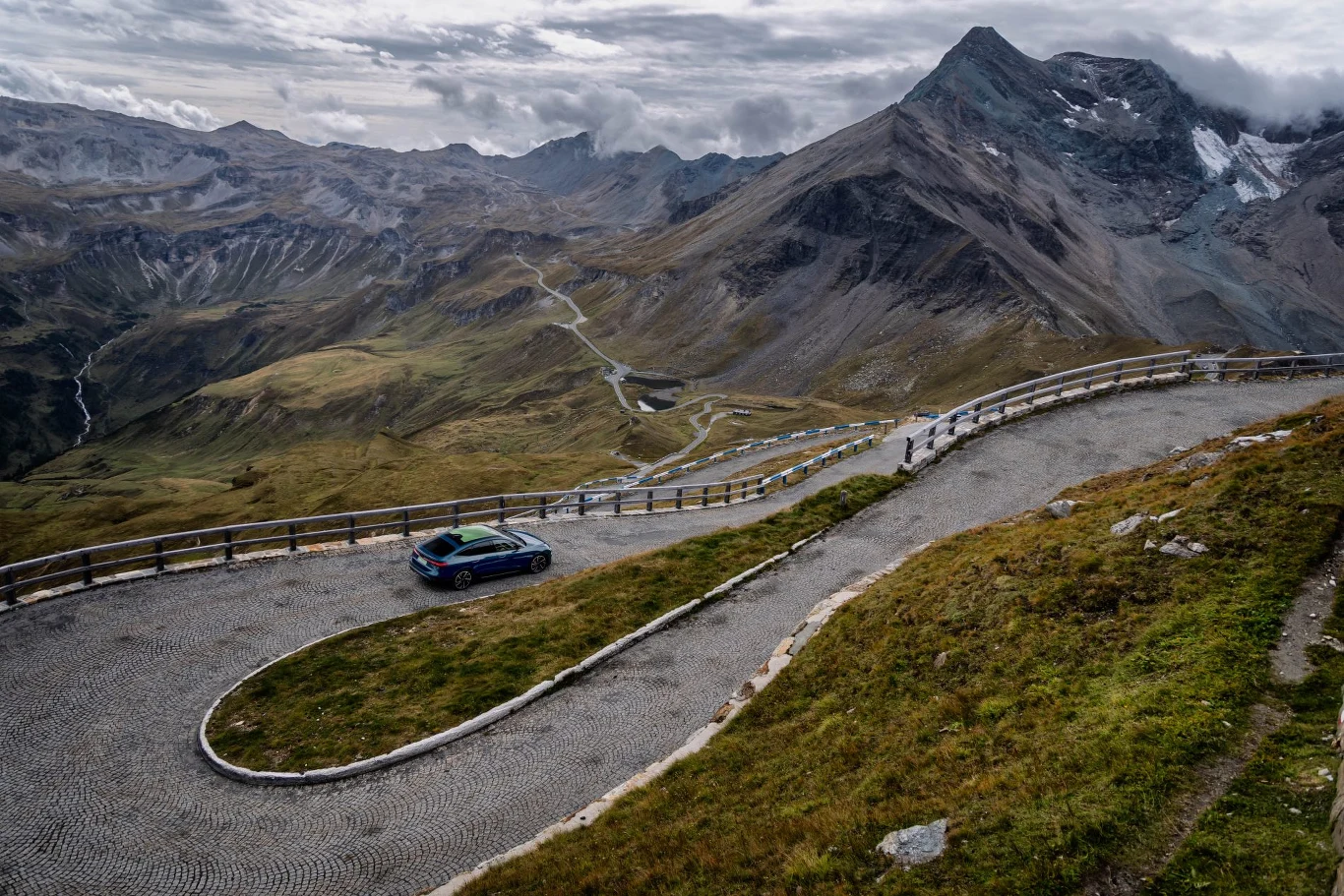 Jedna z najbardziej widowiskowych dróg Europy. Audi S6 e-tron pewnie pokonuje kolejne zakręty nad doliną Pasterze Kręta, brukowana droga górska prowadząca przez zielone zbocza, na zakręcie stoi samochód, w tle majestatyczne szczyty gór oraz chmury nad rozległym krajobrazem.